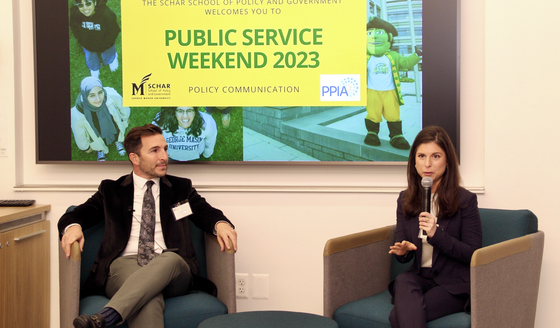 A man in a dark jacket, white shirt, and dark tie sits in a chair next to a woman in a dark outfit holding a microphone.