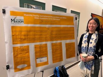 A young woman holding a cup of coffee in front of a research poster wears a black top and a white and blue scarf.