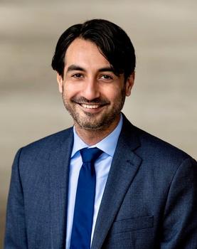 A man with dark hair and a short beard in a suit jacket and blue necktie with a blue shirt smiles at the camera.