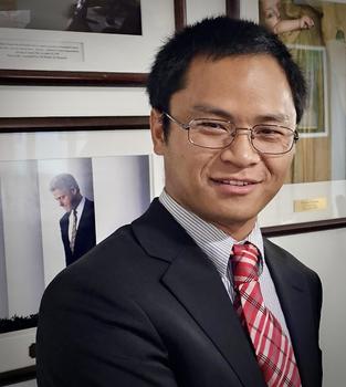 Justin Seo wearing a dark jacket and red necktie smiles at the camera.